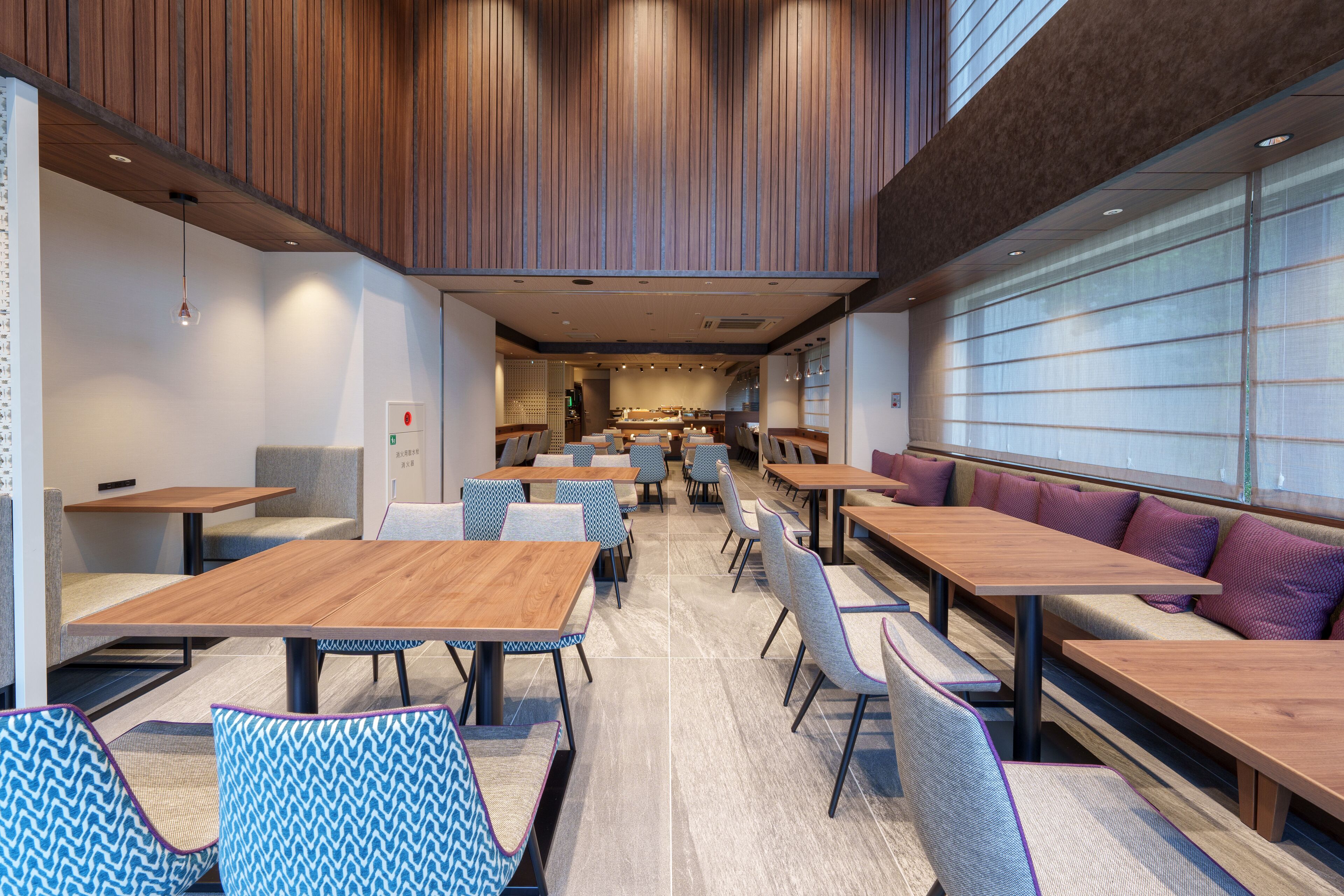 Un espace repas moderne avec des tables et des chaises en bois, un coin salon avec des coussins et, au fond, une vue sur la cuisine.