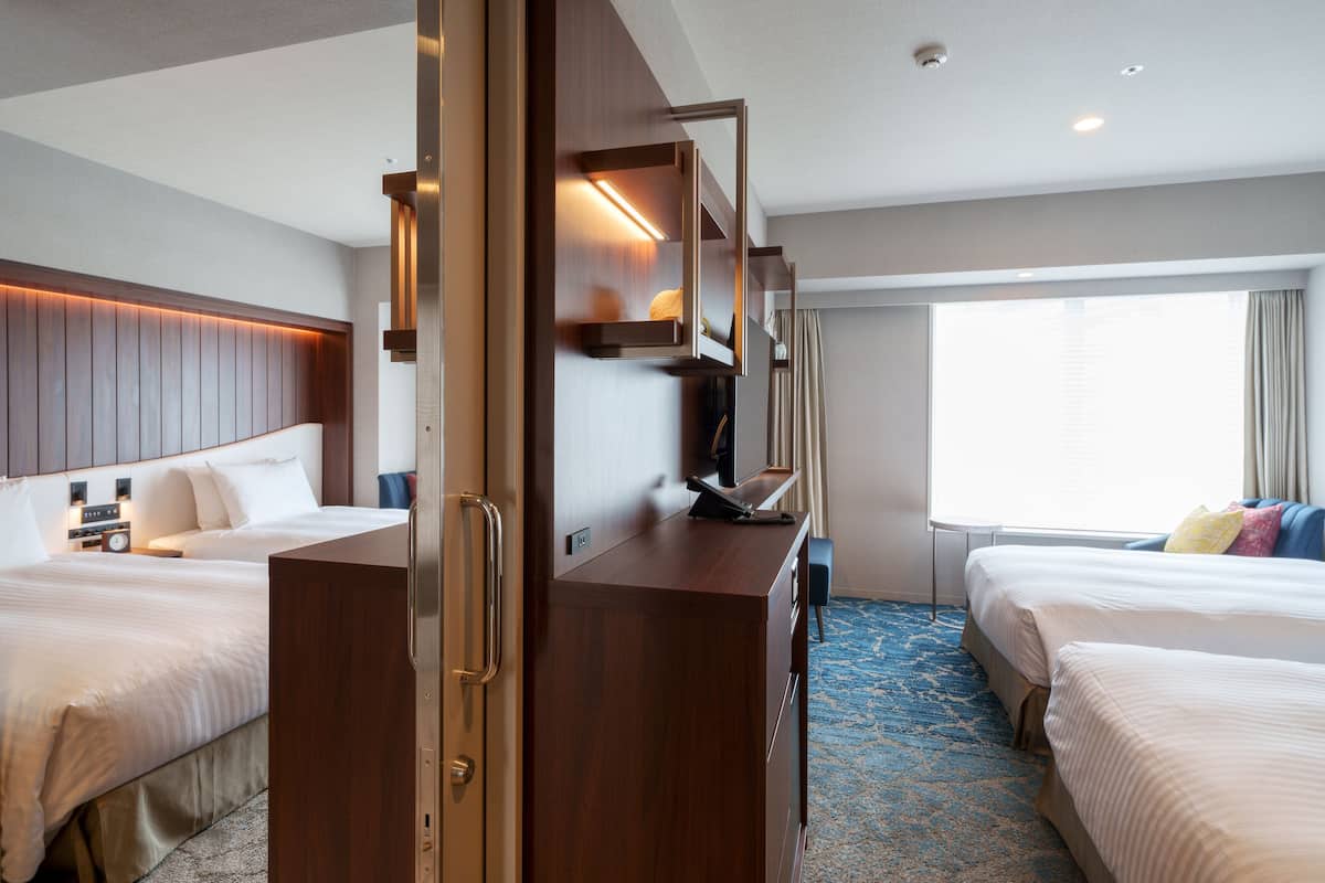 A hotel room with two beds, a wooden desk, a flat-screen TV, and a window with curtains.