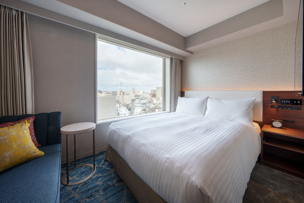 A hotel room with a large bed, a small round table, a sofa with cushions, a nightstand with a clock, and a city view through a large window.