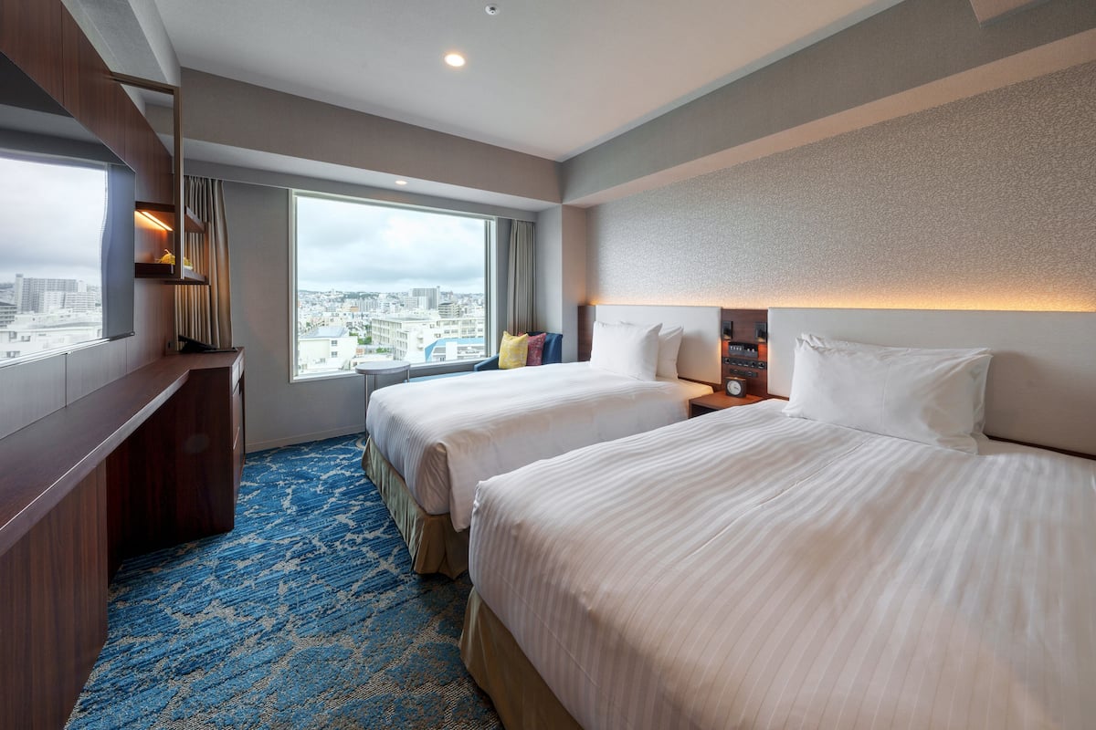 A hotel room with two beds, a large window with a city view, a desk, and a blue carpet.