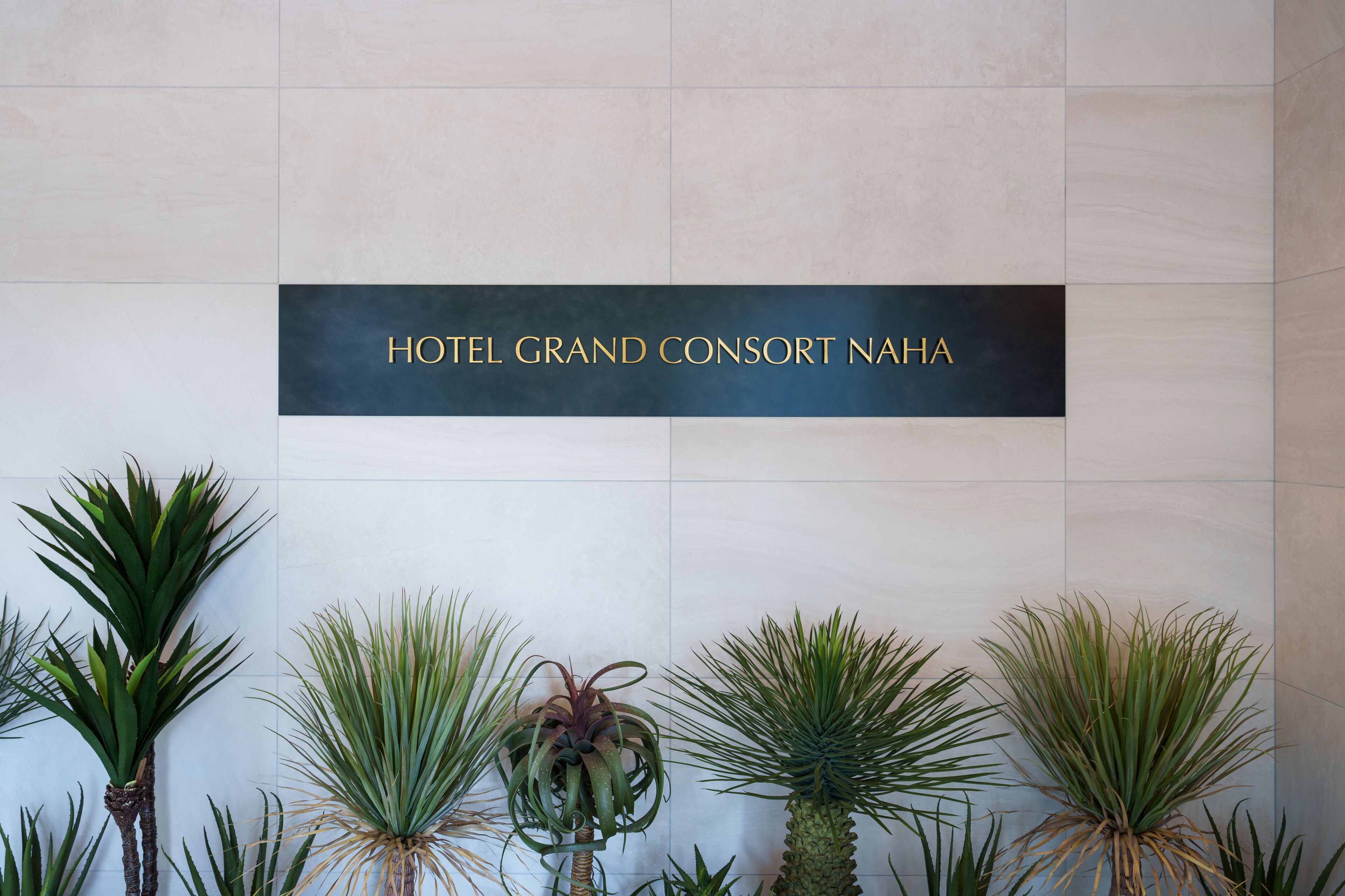 En skylt för HOTEL GRAND CONSORT NAHA, med en vägg klädd i kakel och krukväxter framför.