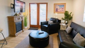 Ett vardagsrum med en öppen spis, en svart lädersoffa, en svart pall och en tv som är monterad på väggen.