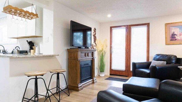 Ett modernt vardagsrum med en svart lädersoffa, ett träbord och en tv som är monterad ovanför en öppen spis. Nära entrén står två barstolar och en krukväxt.