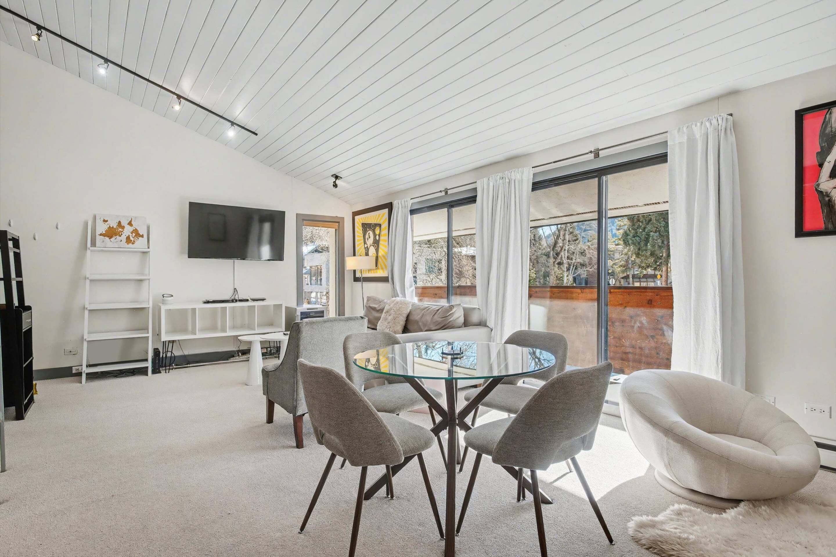 A modern living room with a glass-top coffee table, grey chairs, a sofa, and a flat-screen TV mounted on the wall.