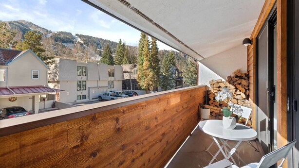 En balkon med gelænder i træ, et hvidt bord og stole. Der ligger et stabel brænde op ad væggen.