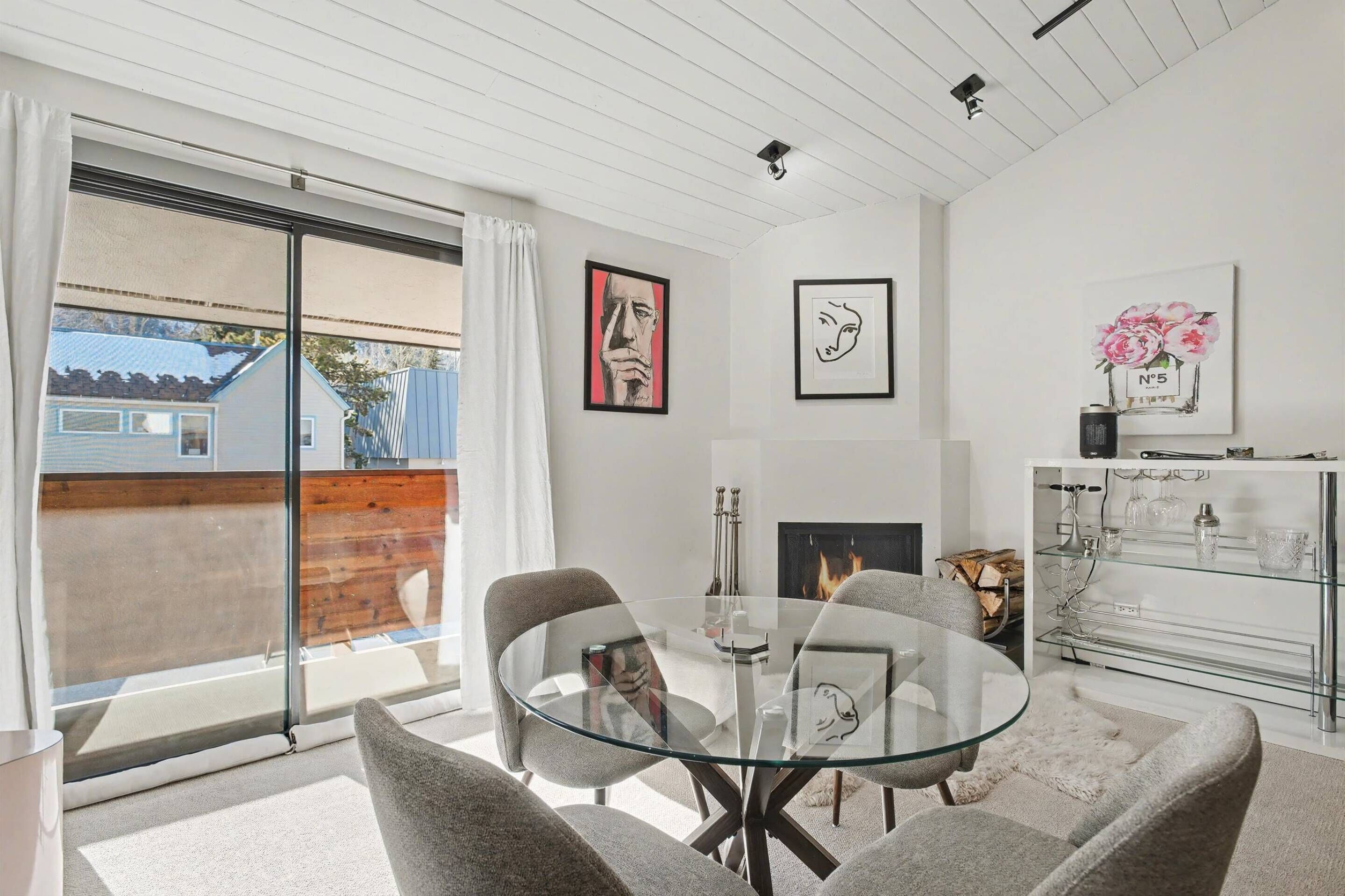A modern living room with a glass dining table, grey chairs, a fireplace, and artwork on the walls.