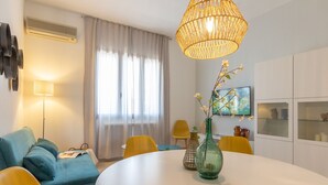 Uma sala de estar moderna com uma mesa redonda, sofá azul, cadeiras amarelas, um lustre de vime e uma TV na parede.
