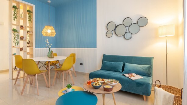 Uma sala de estar moderna com uma parede azul, um sofá azul-esverdeado, cadeiras amarelas e uma área de jantar com uma mesa branca e cadeiras de madeira.