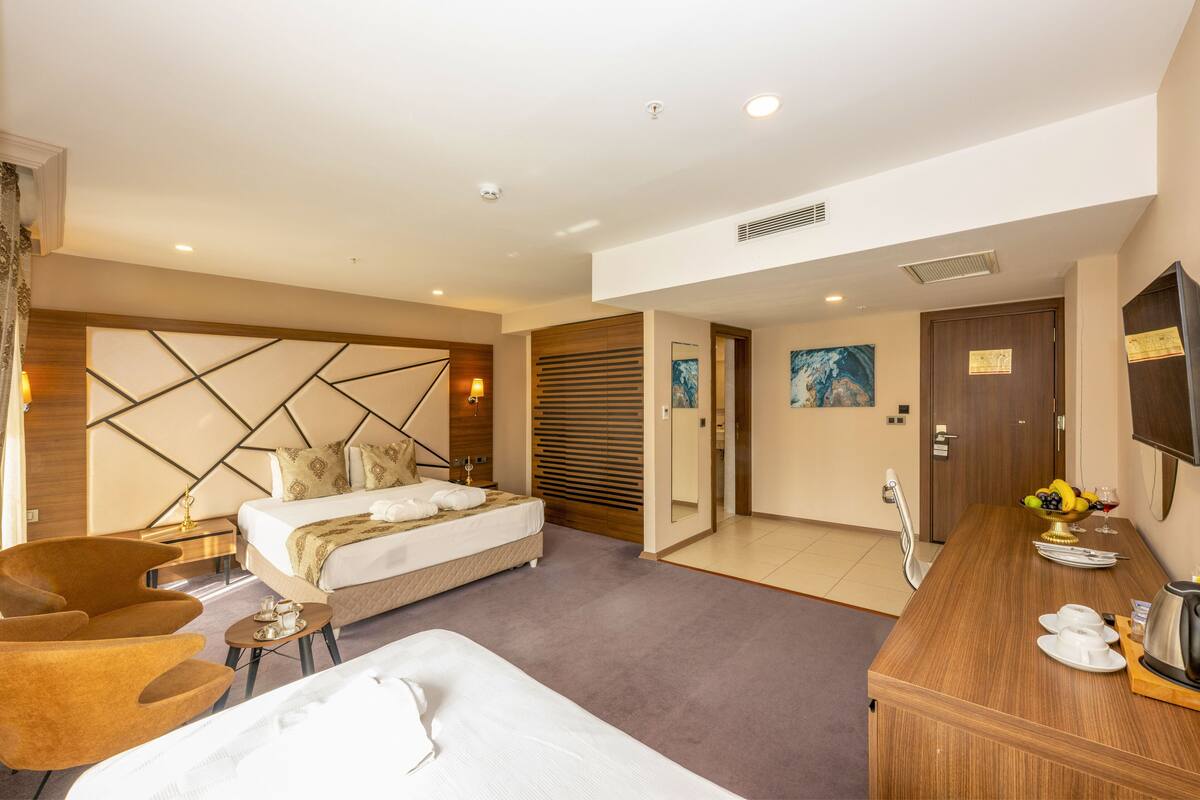 Une chambre d’hôtel moderne avec un grand lit, un coin salon avec un fauteuil, une petite table avec une lampe et un bureau en bois agrémenté d’un panier de fruits et de tasses.