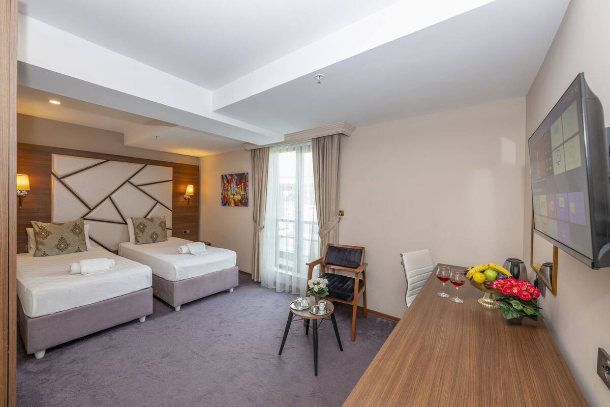 Une chambre d’hôtel avec deux lits, une table à manger agrémentée d’un panier de fruits et de fleurs, et une chaise.