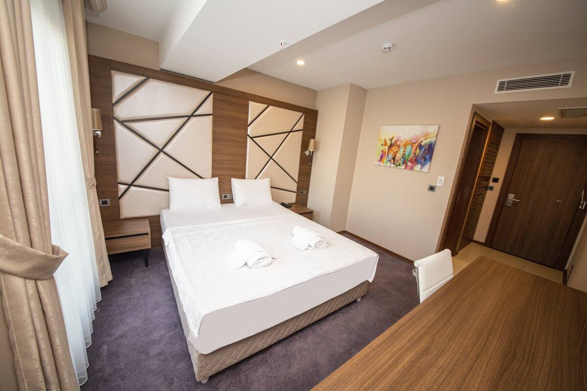 Une chambre d’hôtel avec un grand lit, une table de chevet, une chaise, une tête de lit en bois, une œuvre d’art murale et une porte.