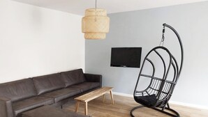 Ein modernes Wohnzimmer mit einem dunkelgrauen Ledersofa, einem Holztisch, einer hängenden Rattan-Leuchte, einem schwarzen Metall-Schaukelstuhl und einem flachen Bildschirm-Fernseher an der Wand.