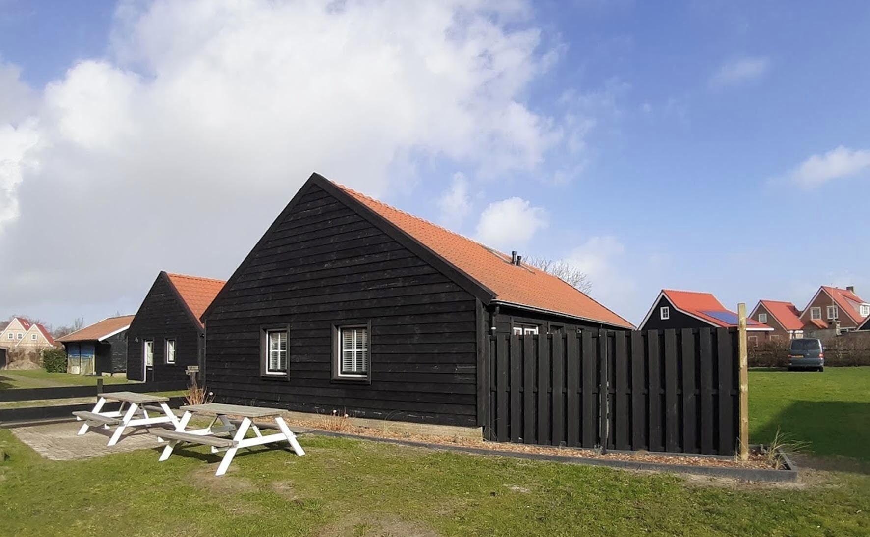 Un bâtiment en bois noir, surmonté d’un toit rouge, entouré d’autres bâtiments similaires, et une table de pique-nique au premier plan.