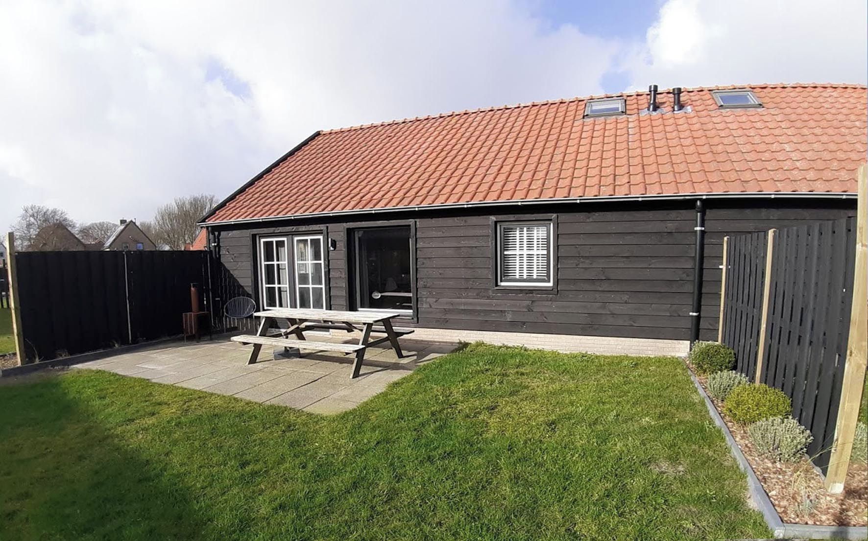 Une maison avec un toit recouvert de tuiles rouges, une façade de couleur gris foncé, une terrasse en bois et une cour arrière verdoyante.