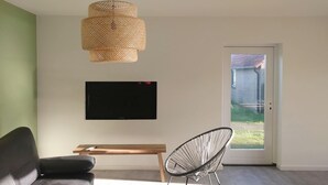 A modern living room with a sofa, a TV mounted on the wall, a woven pendant light, a chair, and a sliding glass door leading to a garden. - Newly renovated apartments on large grounds (Hollum)