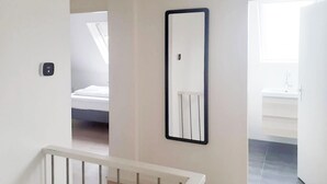 A hallway with a white wall, a black-framed mirror, a visible door, and a glimpse of a bed and bathroom. - Newly renovated apartments on large grounds (Hollum)
