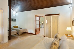 Een kamer met een bed, een nachtkastje, een lamp, een deur en uitzicht op een andere kamer.