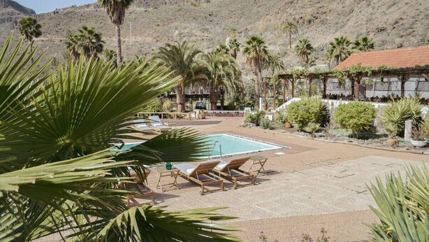 A pool area with lounge chairs, palm trees, and a mountainous backdrop. - Pet-Friendly 'Finca Rural Los Tanquillos' with Mountain View, Pool, Wi-Fi, A/C, Terrace & Garden (San Bartolomé de Tirajana)