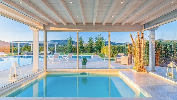A modern indoor pool with clear glass walls, a wooden ceiling, and a view of a landscaped garden and mountains. - Villa Le Terme with Pool, Whirlpool, Garden, Terrace, BBQ, A/C & Wi-Fi (Monsummano Terme PT)