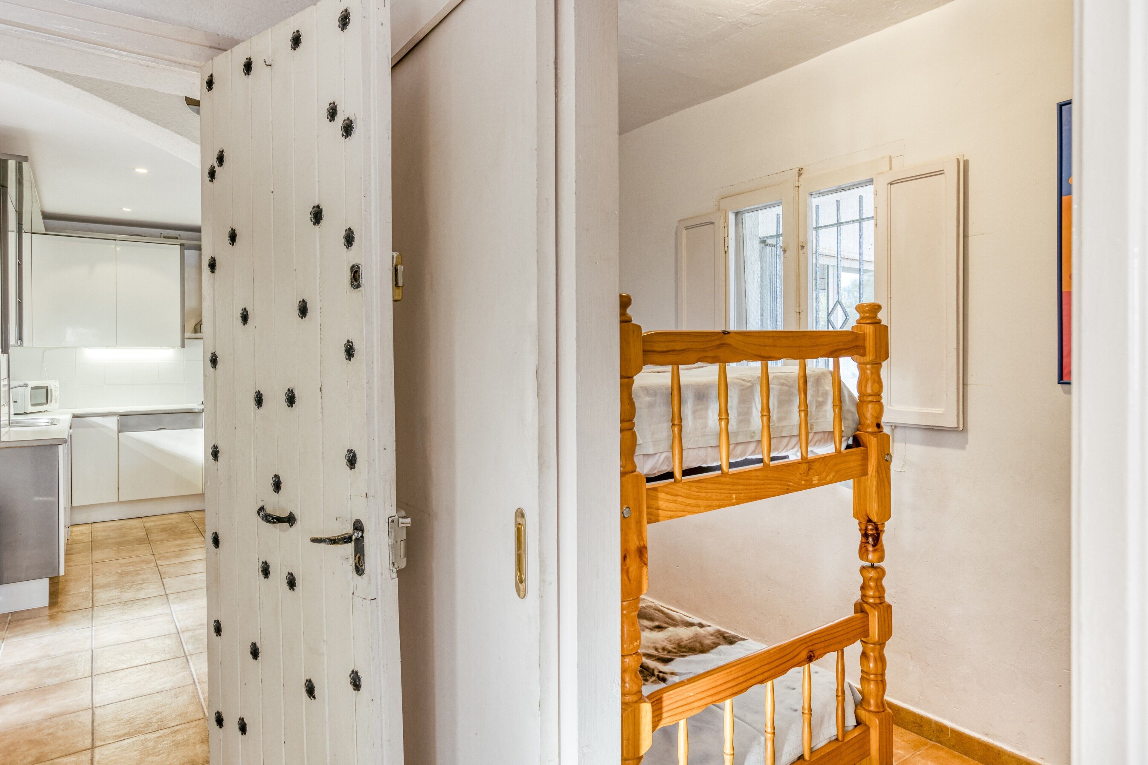 A room with a bunk bed, a white door with decorative elements, and a kitchen area visible through an open door.