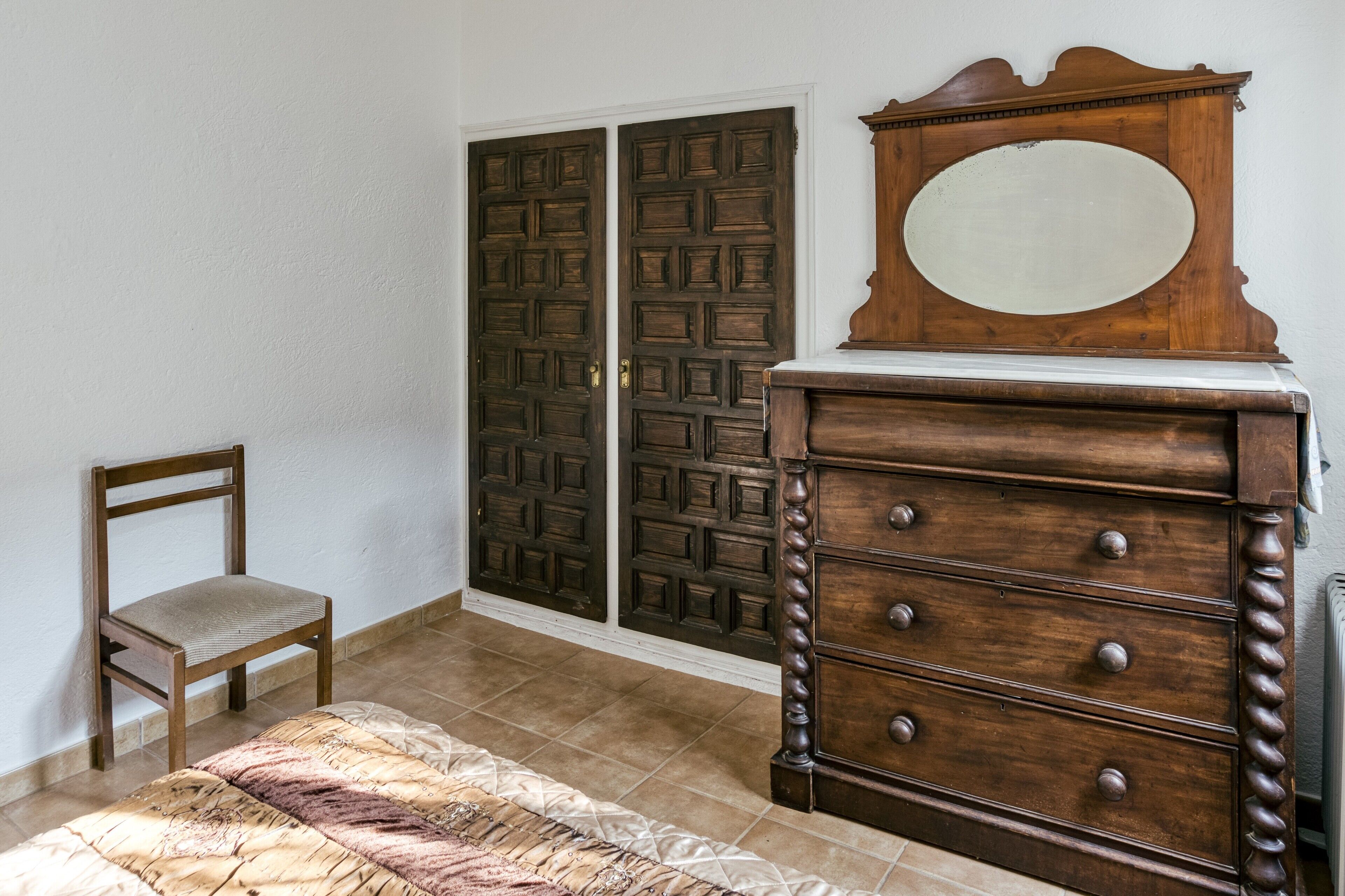 Un dormitorio con una cama, un aparador de madera con cajones, una silla y un doble puerta con diseño geométrico.