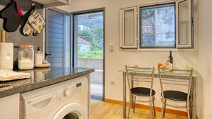 A compact kitchen with a washing machine, a glass table with chairs, and a view through a door to the outside. - Casa Familia Pestana 1, a Home in Madeira (Seixal)