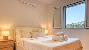 Una cama bien tendida con ropa blanca, una lámpara de noche y una ventana con vistas a una colina.