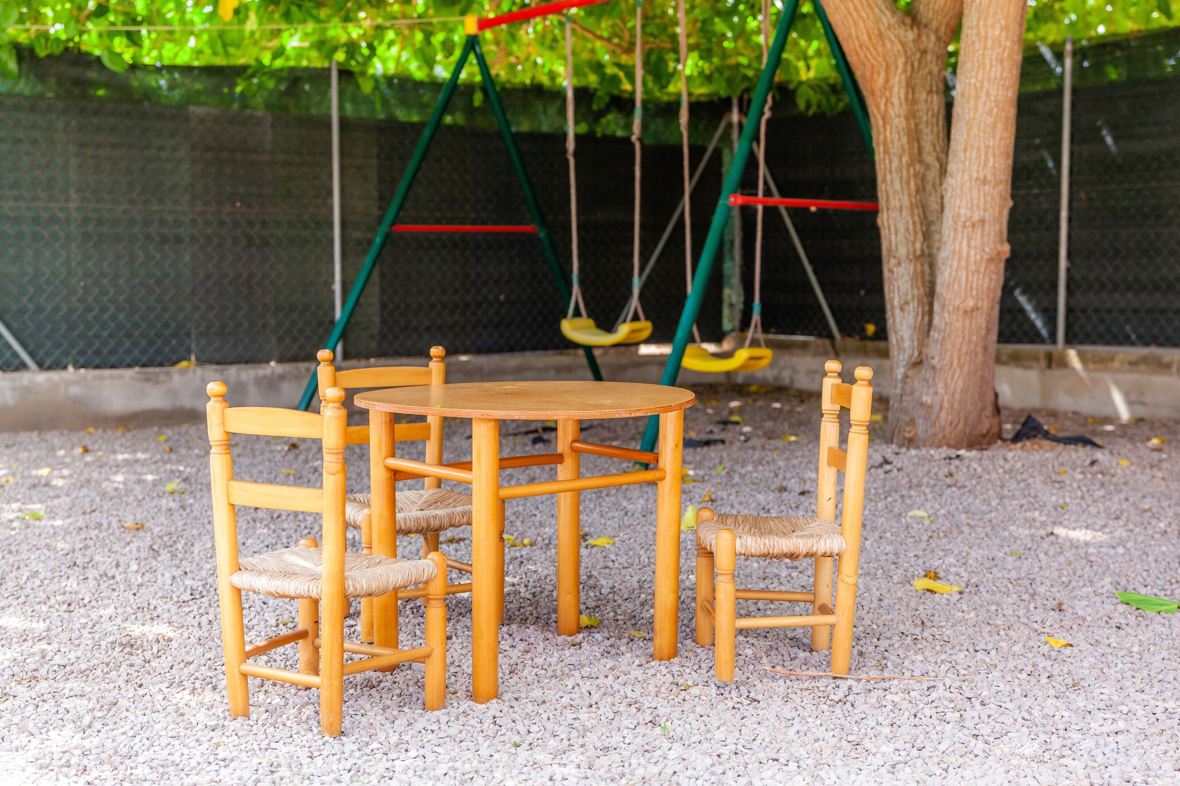 Conjunto de mesa e cadeiras de madeira sobre cascalho, com baloiço e uma árvore ao fundo.