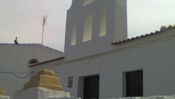 A white building with a cross on top, a balcony with yellow trim, and a small antenna on the roof.