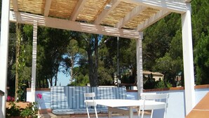 Un espace extérieur aménagé avec une zone salon couverte, comprenant une table et des chaises blanches, un canapé à rayures et une pergola avec des lattes en bambou.