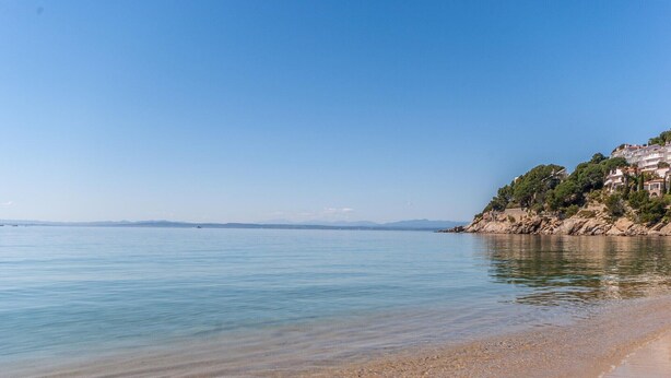 Une plage à l’eau claire, avec des galets et une colline rocheuse parsemée de bâtiments.