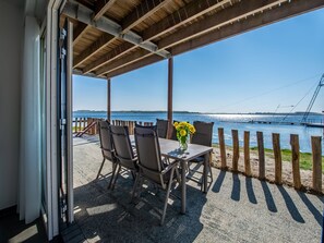 A covered outdoor seating area with a table and chairs, overlooking a body of water with a bridge in the background.
