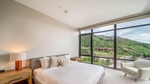 A modern bedroom with a large window, a bed, a nightstand, and a chair. - One Snowmass West 503 (Snowmass)