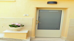 En beige dør med glaspanel og gylden håndtag, en lille hvid postkasse over døren, og en plantekasse med lyserøde blomster til venstre.