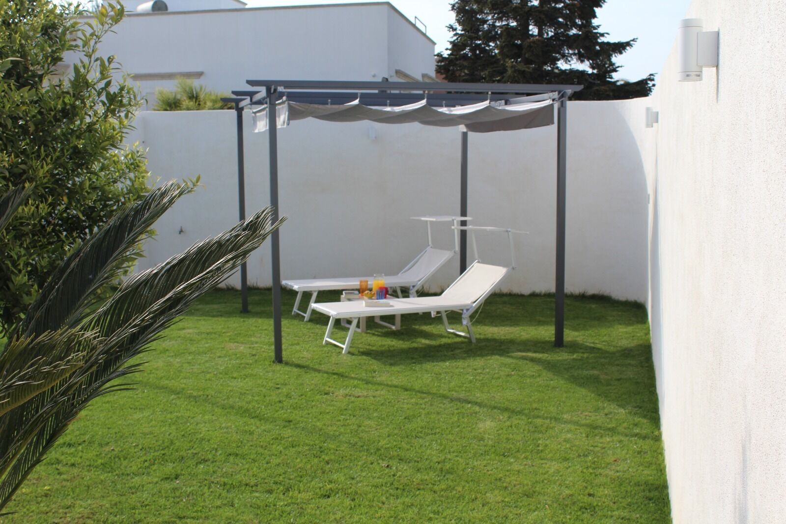 A covered outdoor seating area with two white lounge chairs and a table, surrounded by a white wall and a well-maintained lawn.