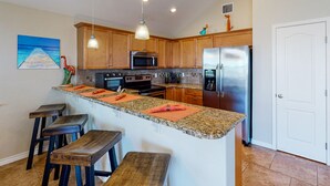 A kitchen with wooden cabinets, a granite countertop, a stainless steel refrigerator, a microwave, and a stove. There are three wooden stools and a painting on the wall.