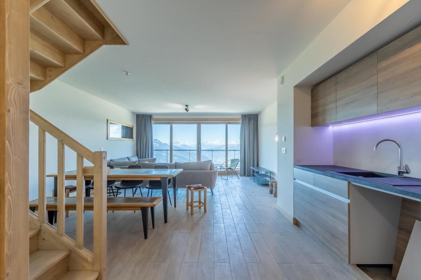 A modern apartment with a wooden staircase, a dining area, a living room with a view, and a kitchen with a built-in island.