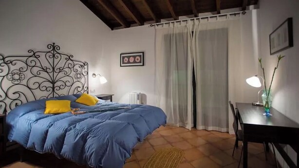 Une chambre à coucher avec une couvre-lit bleue, une tête de lit décorative, une table de chevet avec une lampe, une chaise et une fenêtre avec des rideaux.