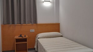 A small, neatly arranged bedroom with a single bed, a bedside table with a lamp, and a window with curtains. - HOTEL AVENIDA (El Morell)