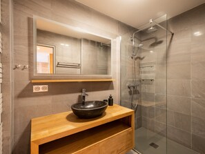 Un baño moderno con un espejo grande, un tocador de madera, un lavamanos de cerámica negra y una ducha con cerramiento de vidrio.