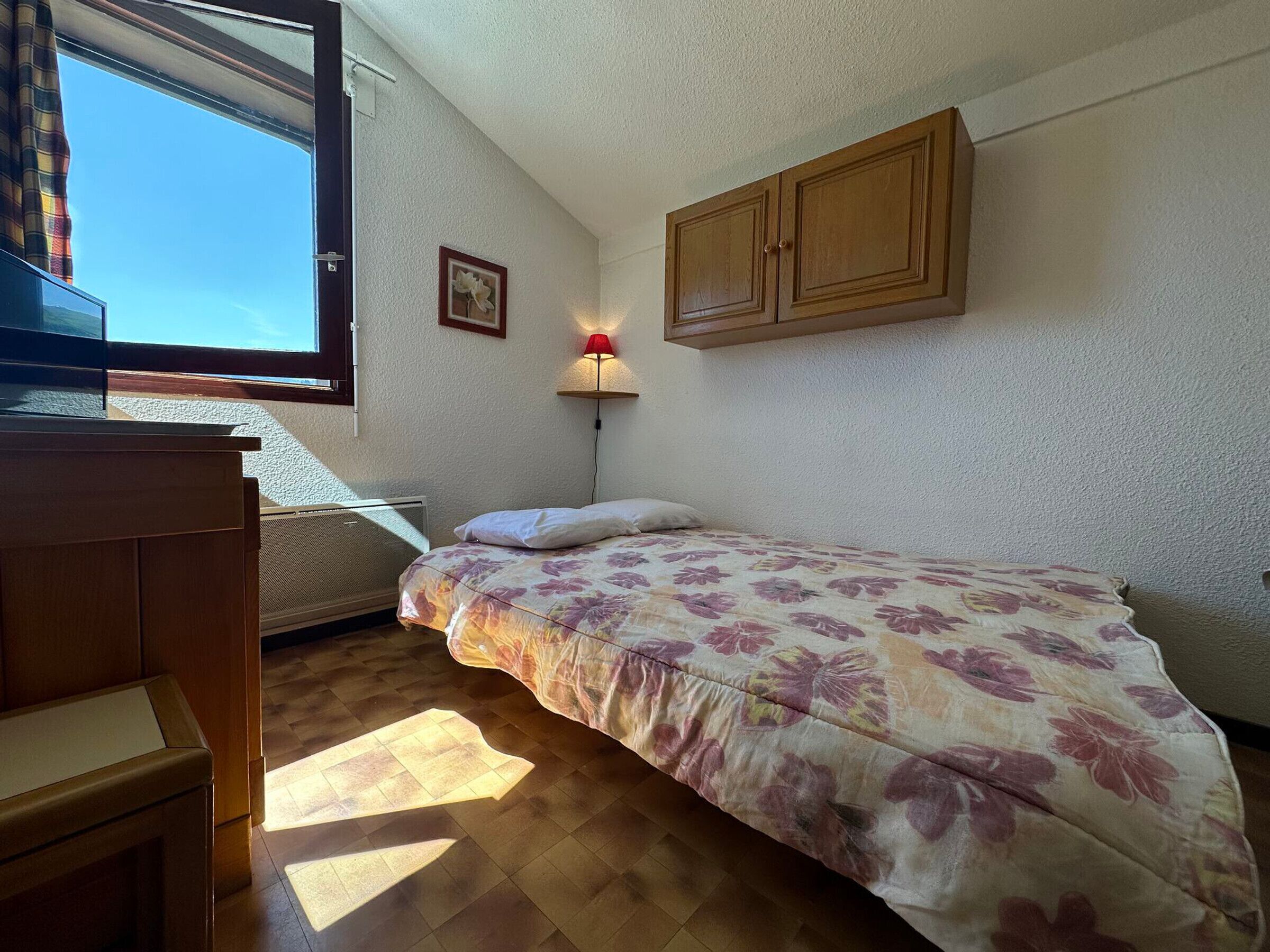 A bedroom with a bed, wooden wardrobe, a desk, a chair, a window with curtains, a wall-mounted lamp, and a framed picture.