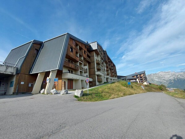A modern multi-story building with a mix of wooden and metal exterior, located near a road with a mountainous backdrop.