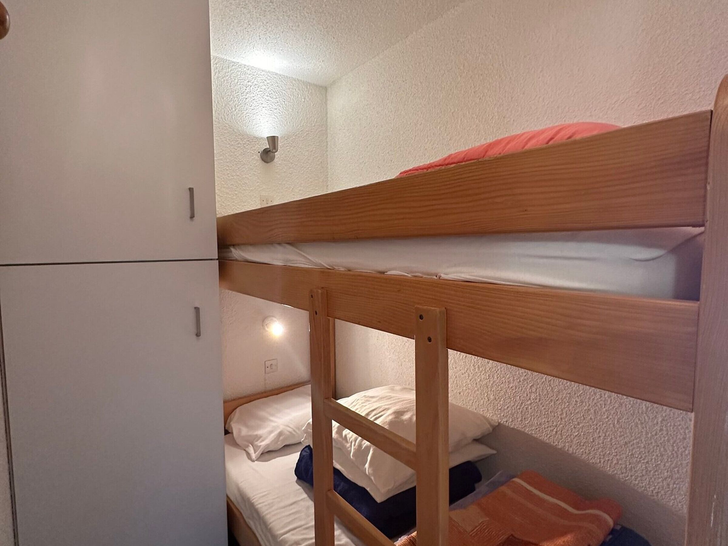 A bunk bed with a wooden frame, white bedding, and a red blanket. There is a white wardrobe with a handle and a light fixture on the wall.