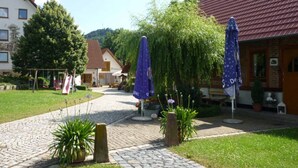 A courtyard with a cobblestone path, potted plants, and two blue umbrellas. - Landhotel Zur Pferdetränke (Schleid)