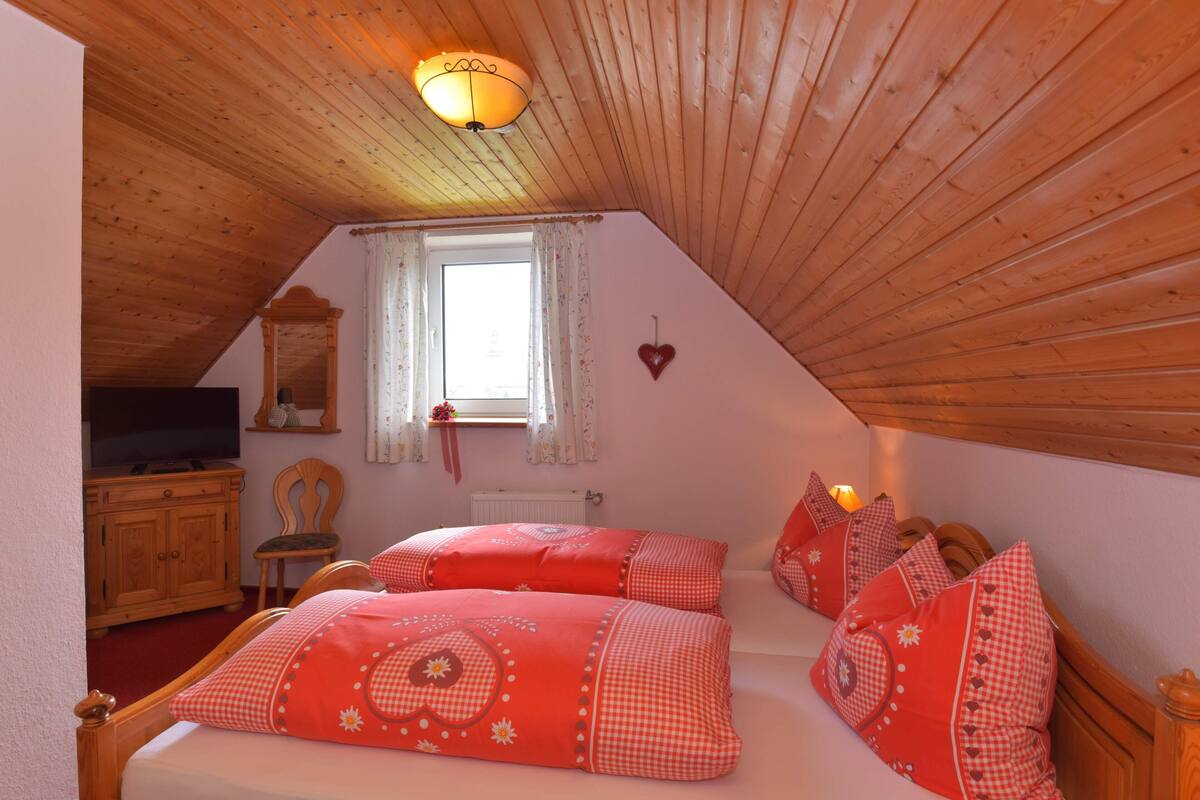 A bedroom with a sloped ceiling, a television on a wooden cabinet, a window with curtains, and a heart-shaped decoration on the wall.