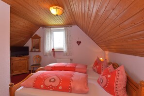 Quarto com teto inclinado, televisão sobre um armário de madeira, janela com cortinas e decoração em formato de coração na parede.
