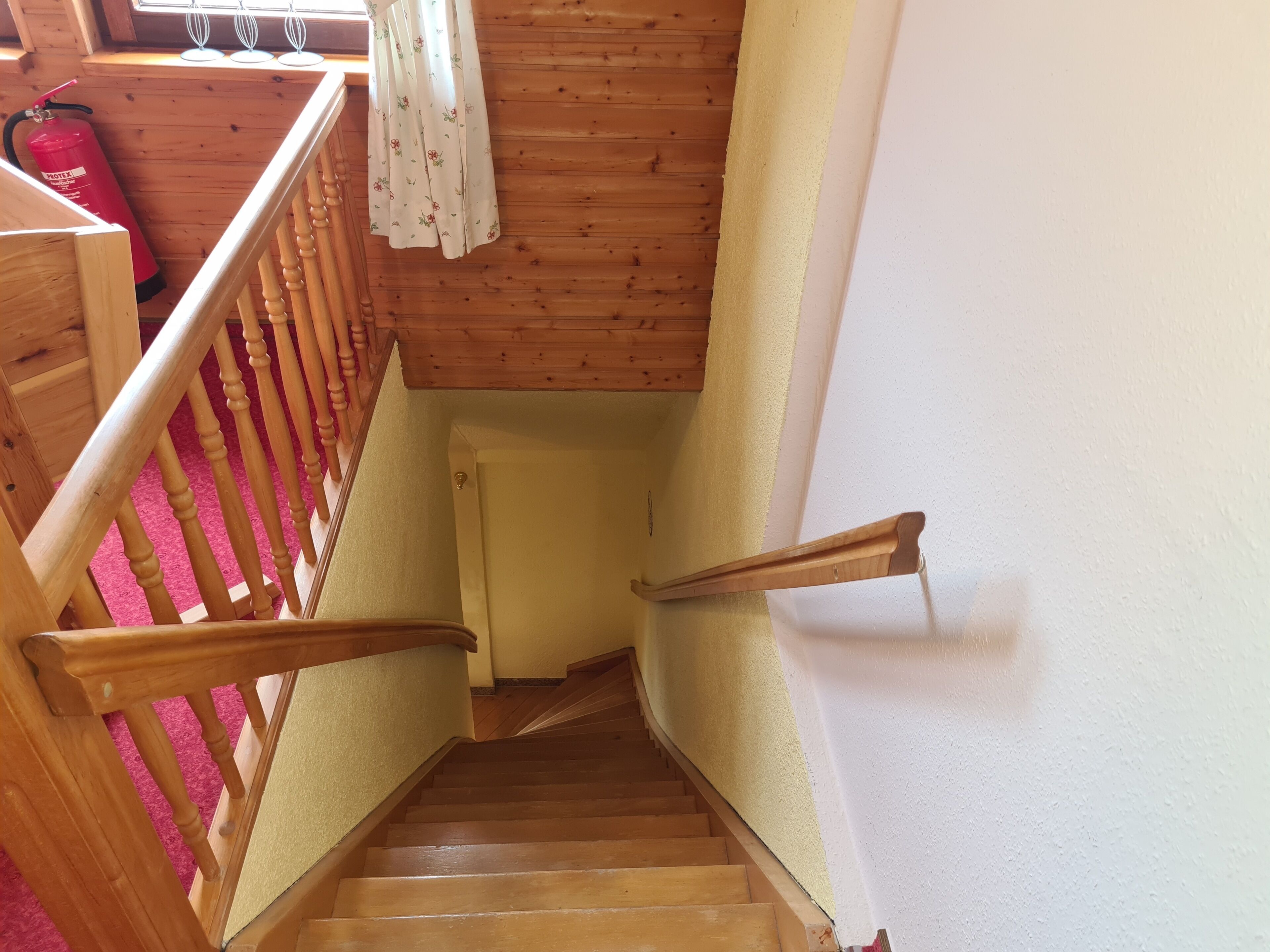 A wooden staircase with a handrail, a fire extinguisher, and a window with curtains.