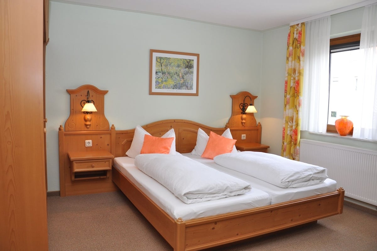 A double bed with white and orange bedding, two bedside tables with lamps, a wooden headboard, a framed picture on the wall, and a window with curtains.