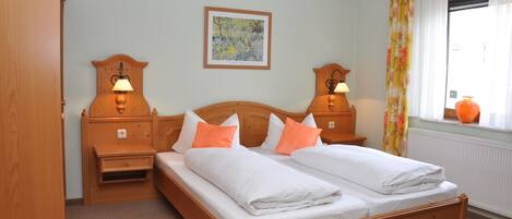 A double bed with white and orange bedding, two bedside tables with lamps, a wooden headboard, a framed picture on the wall, and a window with curtains.