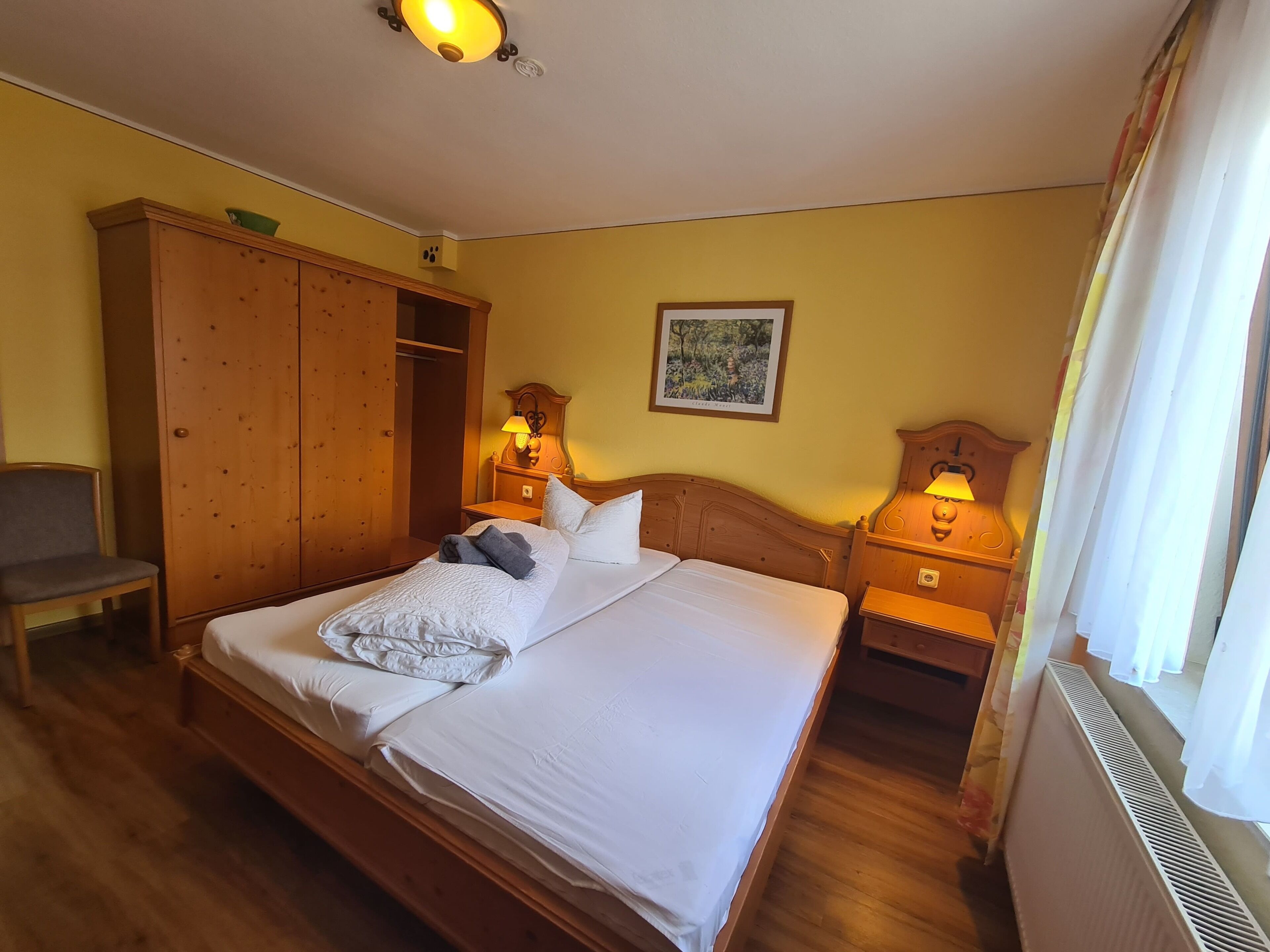Habitación de hotel con una cama de madera, dos mesitas de noche con lámparas, un armario, una silla y un cuadro en la pared.